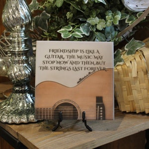 May include: A decorative tile with a brown acoustic guitar and the text "FRIENDSHIP IS LIKE A GUTTAR, THE MUSIC MAY STOP NOW AND THEN, BUT THE STRINGS LAST FOREVER".