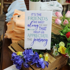 May include: A white sign with the text "IT'S THE FRIENDS WE MEET ALONG THE WAY THAT HELP US APPRECIATE THE JOURNEY" is displayed. The sign is surrounded by flowers, including pink tulips, red roses, and blue and yellow blossoms. Peach-colored shirts are stacked in the background.