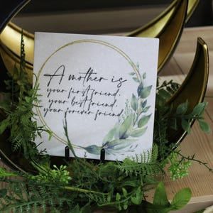 May include: A square decorative tile with the text "A mother is your first friend, your best friend, your forever friend." The tile is surrounded by a gold circle and green leaves. The tile is displayed in a black and gold crescent-shaped holder.