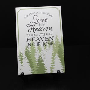 May include: A rectangular sign with the text "Because someone we Love is in Heaven, There's a little bit of Heaven in our Home." The sign has a white background with green pine trees at the bottom. The sign is displayed on a black stand.