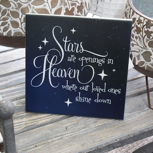 May include: A square, dark blue decorative sign with white script that reads "Stars are openings in Heaven where our loved ones shine down." The sign is decorated with white stars and is placed on a wooden surface.