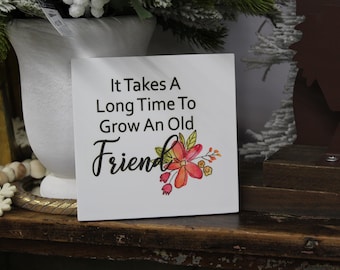 May include: White wooden sign with black text that reads "It Takes A Long Time To Grow An Old Friend". The sign has a watercolor floral design with pink and orange flowers.