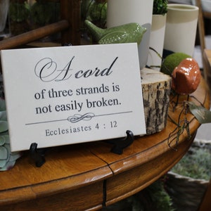 May include: A white canvas sign with black text that reads "A cord of three strands is not easily broken. Ecclesiastes 4:12". The sign is sitting on a wooden table with a small succulent plant and a ceramic bird figurine.