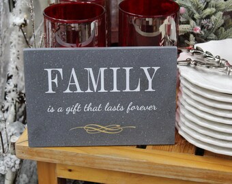 May include: A gray sign with the text "FAMILY is a gift that lasts forever" in white lettering. The sign has a gold swirl design and is sitting on a wooden table.