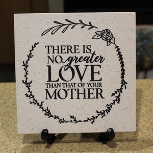 May include: A white ceramic tile with a black floral wreath design and the text "THERE IS NO GREATER LOVE THAN THAT OF YOUR MOTHER".