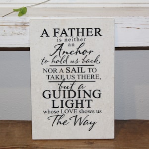 May include: A white plaque with a black inscription that reads "A father is neither an anchor to hold us back nor a sail to take us there, but a guiding light whose love shows us the way."