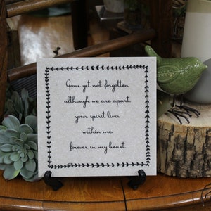 May include: A small, gray, square sign with a black border and a white background. The sign has a black border with a repeating leaf design. The sign reads "Gone yet not forgotten although we are apart. Your spirit lives within me. Forever in my heart."