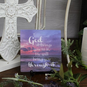 May include: A decorative wooden sign with a purple and pink sunset background and the text "If God brings you to it, He will bring you through it."