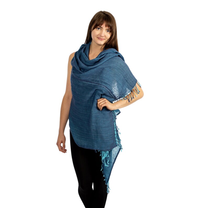 May include: A blue cotton scarf with a fringed edge. The scarf is draped over a person's shoulders and is worn as a shawl.