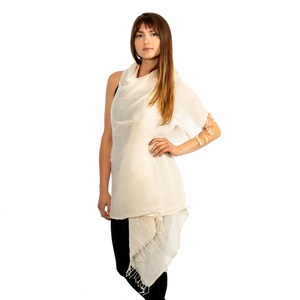 May include: A white, lightweight scarf with a cowl neck and fringed edges. The scarf is draped over a person's shoulders and is worn as a top.