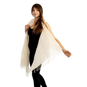 May include: A woman wearing a white cotton scarf with fringe. The scarf is draped over her shoulders and tied at the neck. She is wearing a black sleeveless top and black trousers.