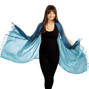 May include: A woman wearing a teal blue scarf with fringe. The scarf is draped over her shoulders and tied at the neck. She is wearing a black tank top and black pants.