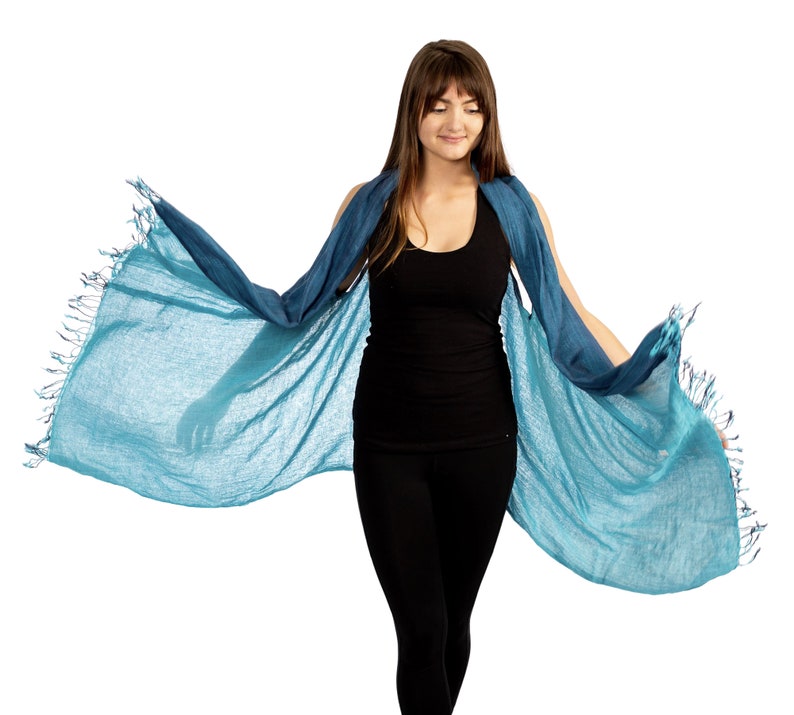 May include: A woman wearing a teal blue scarf with fringe. The scarf is draped over her shoulders and tied at the neck. She is wearing a black tank top and black pants.