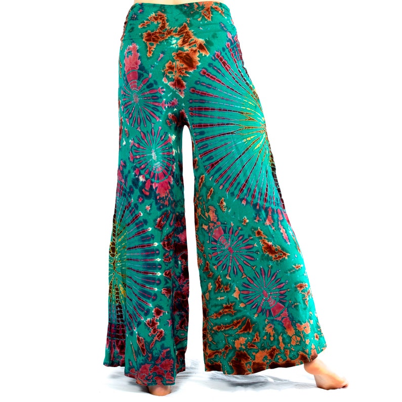 May include: Teal tie-dye wide-leg trousers with a striking pattern. The design includes bursts of pink, brown, and yellow against a teal background. The trousers are made of a light, flowing fabric, ideal for a relaxed style.