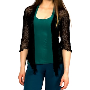 Op de afbeelding: Een zwarte, doorschijnende cardigan met een gerimpelde zoom, gedragen over een teal tanktop en een teal broek.