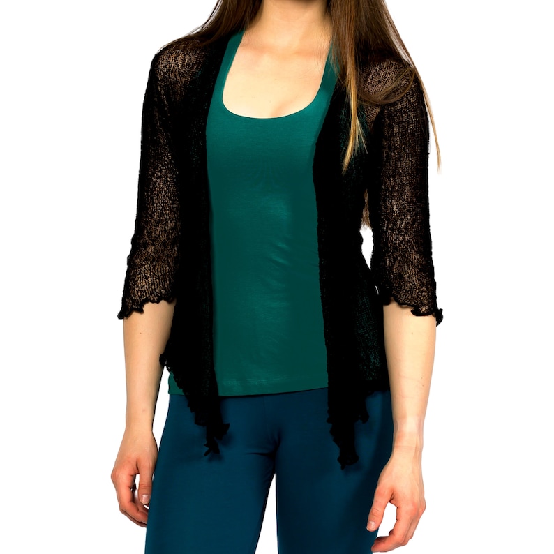 Op de afbeelding: Een zwarte, doorschijnende cardigan met een gerimpelde zoom, gedragen over een teal tanktop en een teal broek.