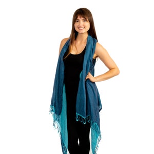 May include: A woman wearing a blue and teal ombre scarf with fringe. The scarf is draped over her shoulders and tied at the neck. She is wearing a black vest top and black trousers.