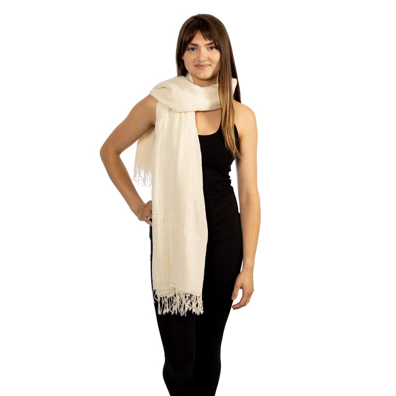 May include: A white linen scarf with fringe edges. The scarf is draped around the neck of a person wearing a black vest top and black trousers.