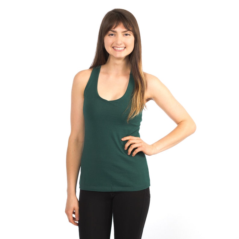 May include: A woman wearing a dark green sleeveless tank top and black trousers. She is smiling and has her hand on her hip.