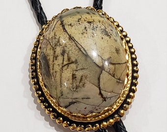 Può includere: Un cravatta a bolo con una grande pietra ovale incastonata in una lunetta color oro. La pietra ha un motivo beige e marrone. Il cordino in pelle nera intrecciata è visibile.