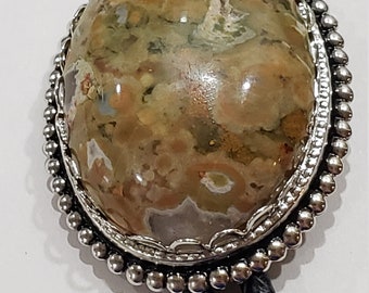 May include: Oval bolo tie with a polished stone pendant. The pendant features a mix of brown, tan, and green hues, set in a silver-toned frame with a beaded edge. Black braided leather cords complete the design.