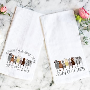 May include: Two white kitchen towels with a goat theme. One towel says "Driving my husband crazy one goat at a time" and the other says "Crazy goat lady".