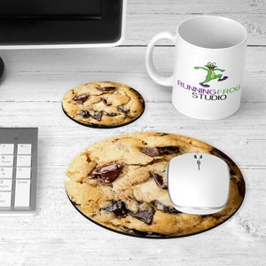 May include: A white ceramic mug with a green frog graphic and the text "Running Frog Studio". The mug is on a white coaster with a chocolate chip cookie on top. There is a computer mouse and a chocolate chip cookie mousepad on a white wooden surface.