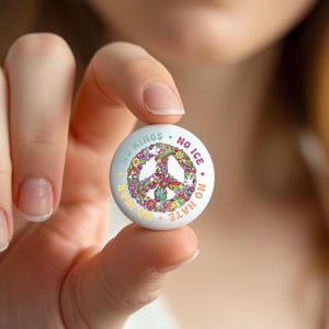 May include: A white button with a peace sign design made of colorful flowers. The button has text that reads "NO KINGS NO WAR NO ICE NO HATE" in a circular pattern around the peace sign.