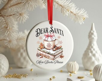 May include: White ceramic Christmas ornament with a red ribbon. The ornament features the text "Dear Santa" and "More Books Please". A graphic of a mug with whipped cream, gingerbread man, and books is also on the ornament.