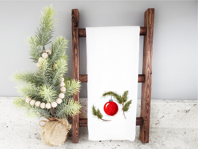 May include: White kitchen towel with a red Christmas ornament and green pine branches embroidered on it. The towel is hanging on a wooden ladder.