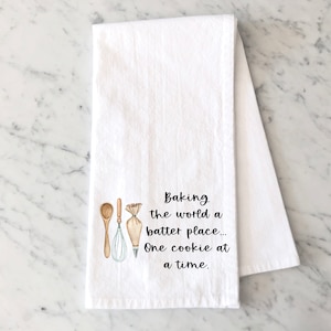 May include: White kitchen towel with a brown wooden spoon, a whisk, and a piping bag. The text reads "Baking the world a batter place... One cookie at a time."