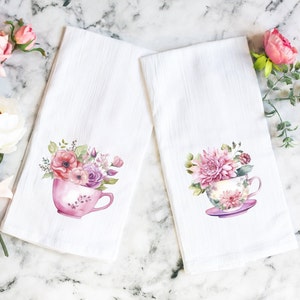 May include: Two white tea towels with floral designs. Each towel features a pink teacup filled with pink and purple flowers. The teacups are on a white background.