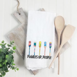 May include: White kitchen towel with a colorful illustration of six paddles and the text "paddles up, people".