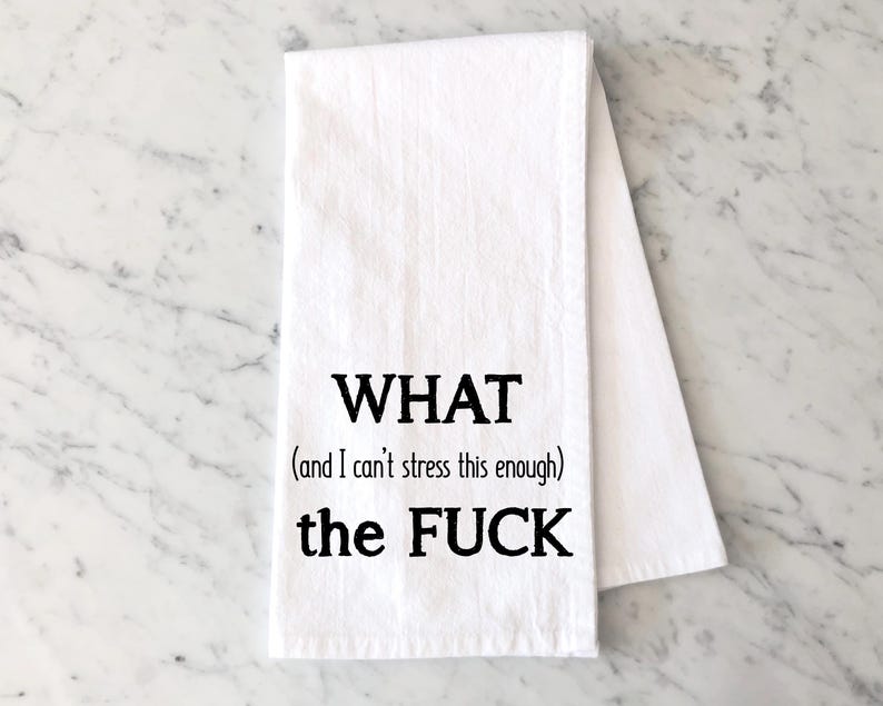 May include: White kitchen towel with black text that reads "WHAT (and I can't stress this enough) the FUCK". The towel is folded and lies on a marble surface.