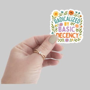 May include: A colorful sticker with the text "RADICALIZED BY BASIC DECENCY" surrounded by floral illustrations. The sticker is held in a hand with a gold ring.