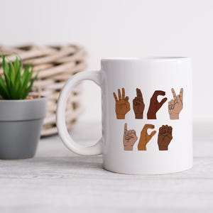 K&ouml;nnte beinhalten: Wei&szlig;e Keramik-Tasse mit dem Wort "LOVE" in amerikanischer Geb&auml;rdensprache, dargestellt mit H&auml;nden in verschiedenen Hautt&ouml;nen. Die Tasse hat einen wei&szlig;en Henkel. Die Tasse steht auf einer hellen Oberfl&auml;che, mit einer kleinen Pflanze und einem Weidenkorb im Hintergrund.