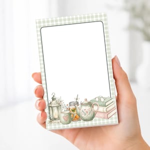 May include: A rectangular notepad featuring a light green and white gingham border. The bottom section displays a watercolor illustration of a lantern, a heart-adorned mug, a glass jar, a teapot, and a stack of books with glasses on top.