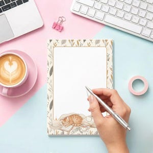 May include: A rectangular notepad featuring a white center bordered by a pattern of brown and beige leaves. The bottom of the page displays an illustration of a sleeping orange cat, a lit candle, a small book, and a cup of tea on a soft blanket.