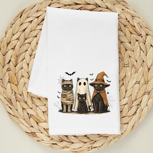 May include: White Halloween-themed kitchen towel featuring a cartoon illustration of four black cats dressed as a mummy, ghost, witch, and a small black cat. Bats and spiderwebs are also included in the design. The towel is folded on a woven, circular placemat.