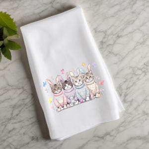 May include: Four cats wearing pastel colored sweaters with bunny ears. The cats are sitting in a row and have different colored sweaters. The background is white.