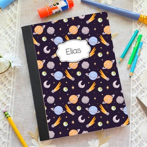 May include: A rectangular notebook with a dark purple cover featuring a repeating pattern of planets, comets, stars, and moons. A white label with a decorative border is centered on the front cover and contains the printed name Elias.