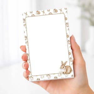 May include: A notepad with a white interior and a border featuring a repeating pattern of watercolor bunnies. A single bunny illustration is in the lower right corner. The notepad is held in a hand.