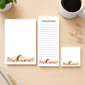 May include: A set of three white paper notepads featuring a watercolor illustration of a hedgehog surrounded by autumn leaves at the bottom. The tall notepad includes horizontal lines and the text Prickly Priorities at the top.