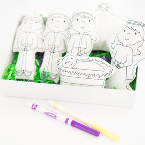 May include: A set of white felt Christmas characters in a white box with green and purple shredded paper. The characters include Mary, Joseph, a shepherd, an angel, and baby Jesus in a manger. Two markers are included for coloring the characters.