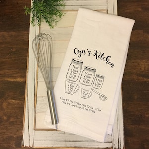 May include: A white kitchen towel with the text "Cyn's Kitchen" printed in black. The towel features a graphic of various sized jars with measurements in gallons, quarts, pints, cups, and ounces. The text "1 Tbsp 1/2 Tbsp 1/4 Tbsp 1 Tsp 1/2 Tsp 1/4 Tsp" and "1 Cup 3/4 Cup 1/2 Cup 1/3 Cup 1/4 Cup" is printed below the jars.
