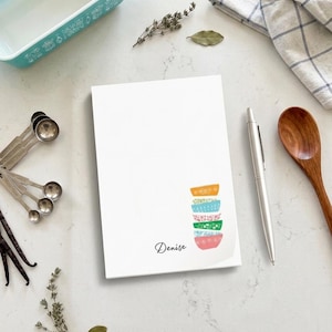 May include: A white notepad with a stack of colorful bowls and the name "Denise" in script. A silver pen, wooden spoon, measuring spoons, and a turquoise baking dish are also visible, suggesting a kitchen theme.