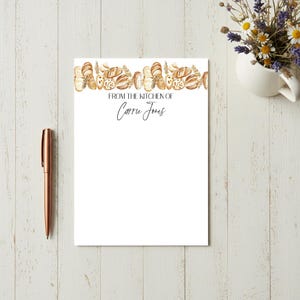 May include: White notepad with a watercolor illustration of various breads along the top edge. The text "FROM THE KITCHEN OF Carrie Jones" is printed in black script. A rose gold pen rests on the left side of the notepad.
