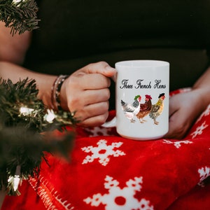 May include: White ceramic mug with a black design of three hens wearing berets and the text "Three French Hens". The mug is being held by a person wearing a black shirt and a red blanket with white snowflakes.
