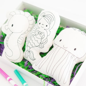 May include: A set of three white plush toys in a box with green and purple shredded paper. The toys are a seahorse, a mermaid, and a jellyfish. The toys are all outlined in black and ready to be colored.