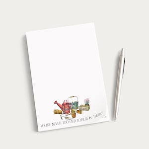 May include: A white notepad with a watercolor illustration of gardening tools, including a red watering can, trowel, and seed packets. The text at the bottom reads, "You're never too old to play in the dirt." A silver pen rests on the side.